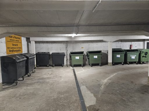 Objekt Nr.28 - Schwenningen Reihe von Müllcontainern in einer Tiefgarage, graue Wände und künstliche Beleuchtung.