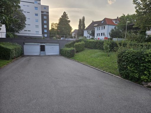 Objekt Nr.6 - VS-Schwenningen Garagenzufahrt zwischen Hecken, im Hintergrund Wohngebäude und leicht bewölkter Himmel.