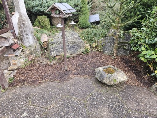 Objekt Nr.1 Nachher - VS-Marbach Ein Gartenbereich mit einem Stein, Pflanzen und einem Vogelhaus im Hintergrund.