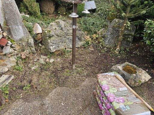 Objekt Nr.1 Vorher - VS-Marbach Ein unebener Gartenboden mit Steinen, einer Lampe und einem bepflanzbaren Kasten.